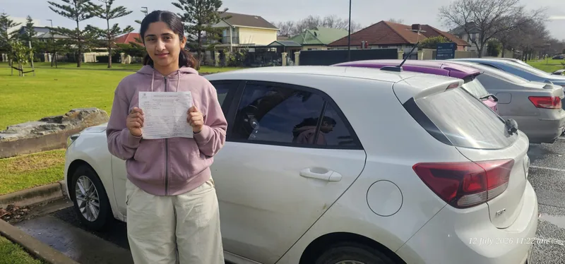 Learner in a pink hoodie holding a certificate beside a white hatchback